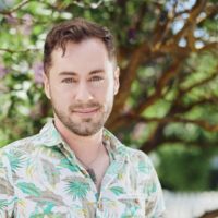 Headshot of an individual with short brown hair, close shaved beard, and floral (with palm trees), button up shirt.