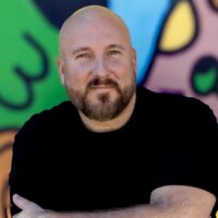 Headshot of a bearded individual wearing a black t-shirt, arms crossed. Man is in front of a brightly colored wall