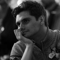 Black and white headshot of an individual, leaning chin on palm. Man is smiling and wears a sweater with a small flower pinned on chest