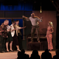 Image of an individual wearing sunglasses, standing on a piano bench, with a crowd of five other individuals around.