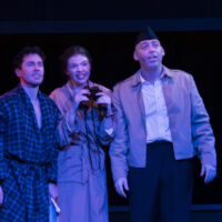 Three individuals in a stage production of Merrily We Roll Along. Middle person is holding binoculars.