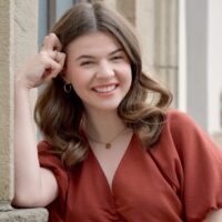 Headshot of a smiling individual, right elbow leaning on edge of building. Right hand pressed against their temple.