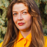 Headshot of an individual with long hair, wearing a yellow shirt, buttoned to the neck.