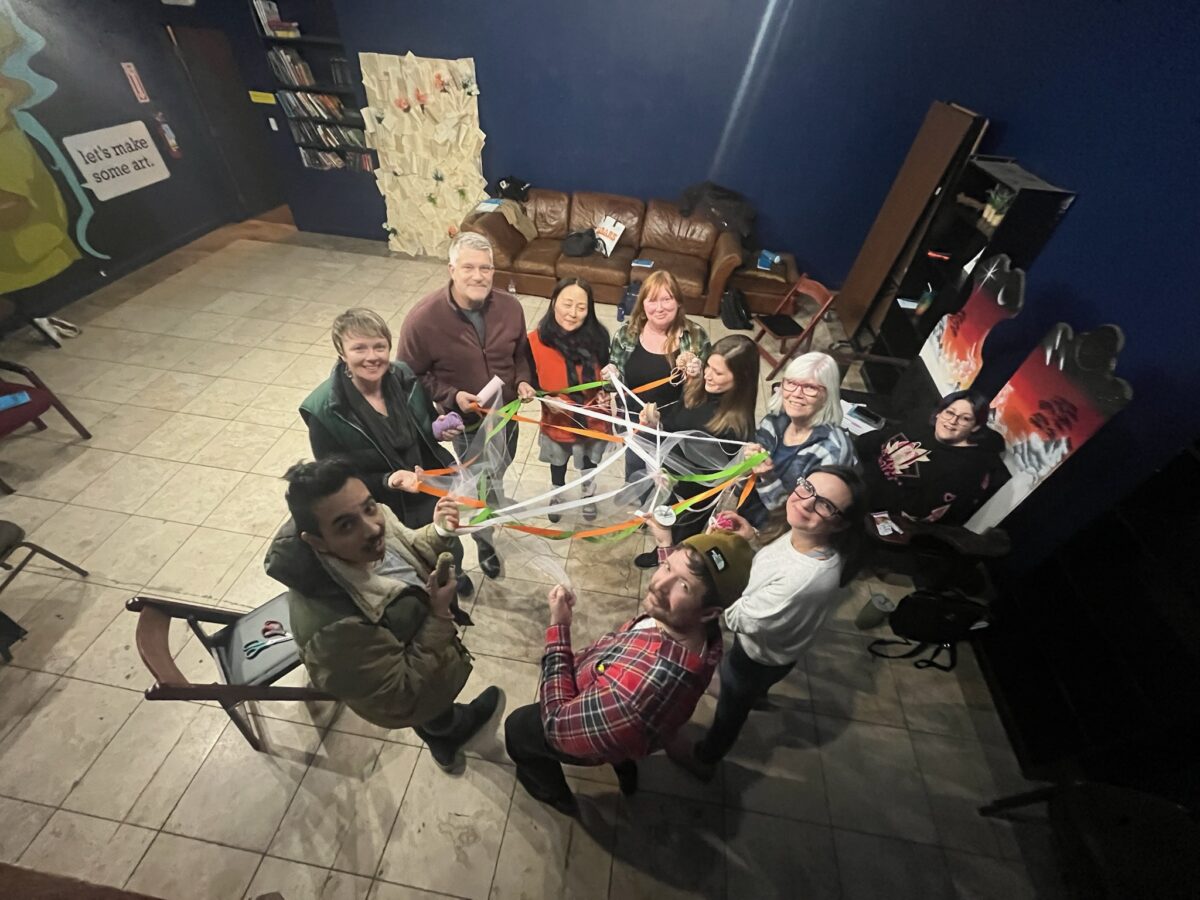 Image of nine people in a circle, holding onto tangled string, looking into the air towards the photographer.